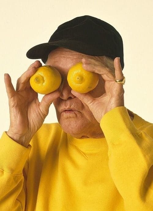 An elderly person in a yellow sweater and black cap  playfully holding two lemons over their eyes against a plain background.