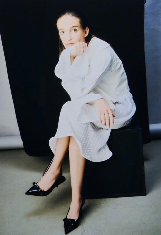 A woman sitting against a dark background, dressed in a white outfit with black pointed shoes,  gazing thoughtfully at the camera.