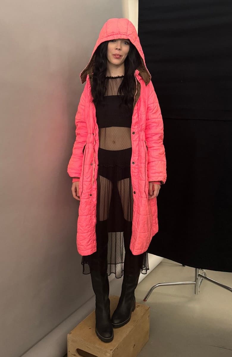 Model wearing a bright pink puffer coat with a hood over a black crop top and leather pants,  standing in front of a plain backdrop.