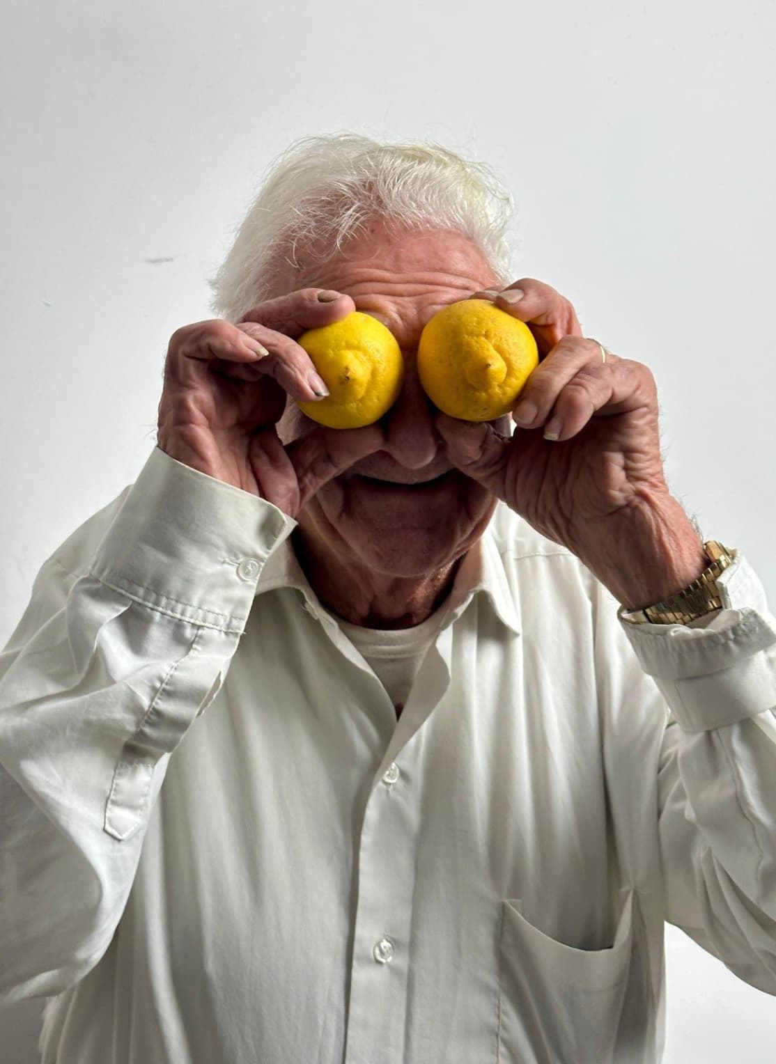 An elderly person with white hair and a white shirt  holding two lemons over their eyes and smiling against a light background.