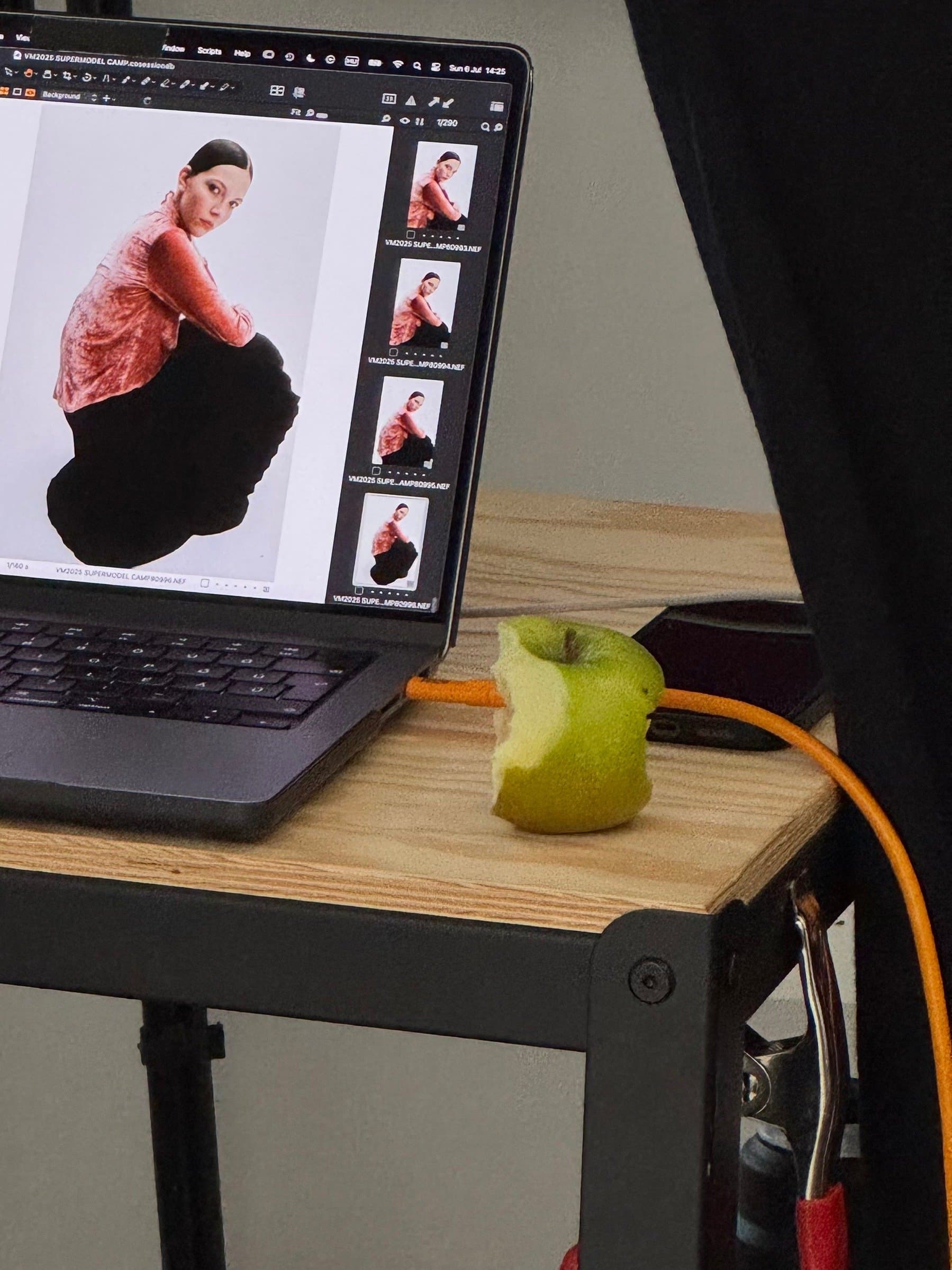 Behind-the-scenes photo showing a laptop displaying a photoshoot image of a model in a red and black outfit,  next to an apple and camera equipment on a studio table.
